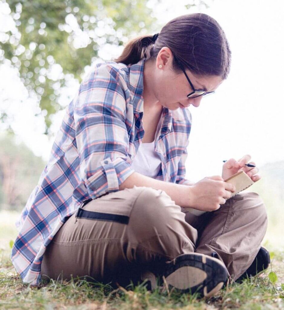Une cliente en introspection est assise en tailleur sur l’herbe et note ses observations sur un calepin.