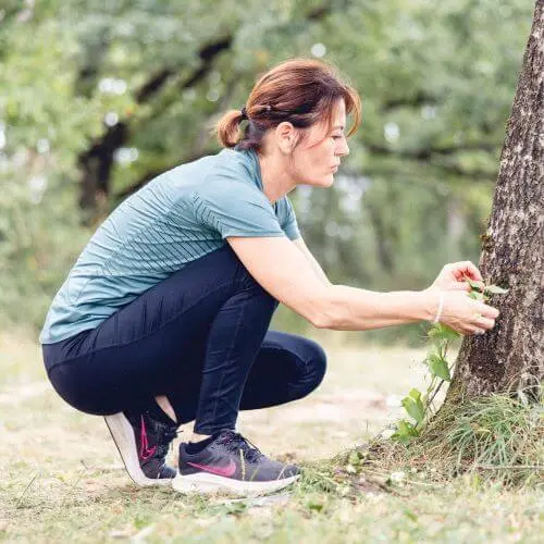 Dans une forêt jurassienne, une femme est accroupie et touche une plante avec ses deux mains.