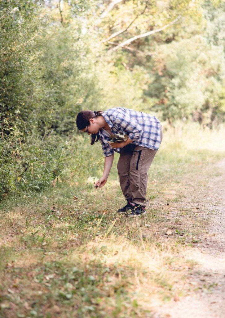Près d’un sentier en forêt, une femme cultive son lien avec la nature en touchant une fleur avec sa main droite.