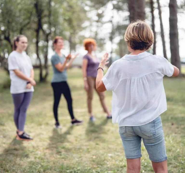 Dans une belle forêt du Jura, la formatrice utilise ses mains pour expliquer les principes de l’écopsychologie à un groupe de participants qui se tient debout devant elle.