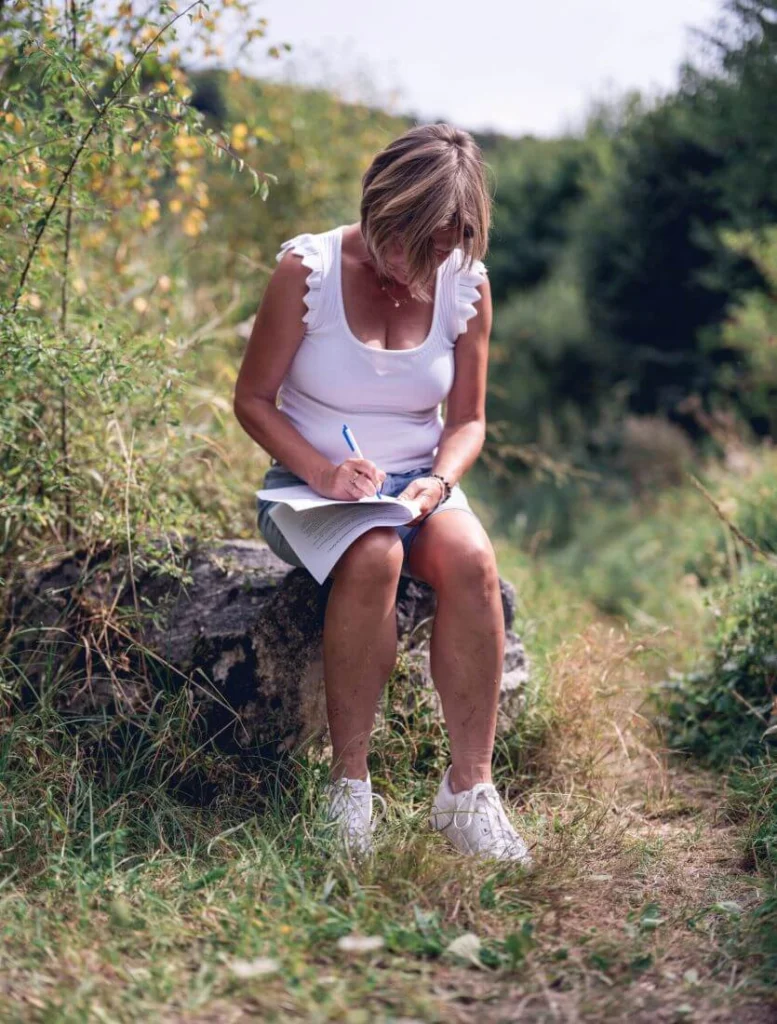 Tout près d’un petit chemin de nature, Ilse Geusens est assise sur un gros caillou et est en train de prendre des notes sur son fascicule de formation.