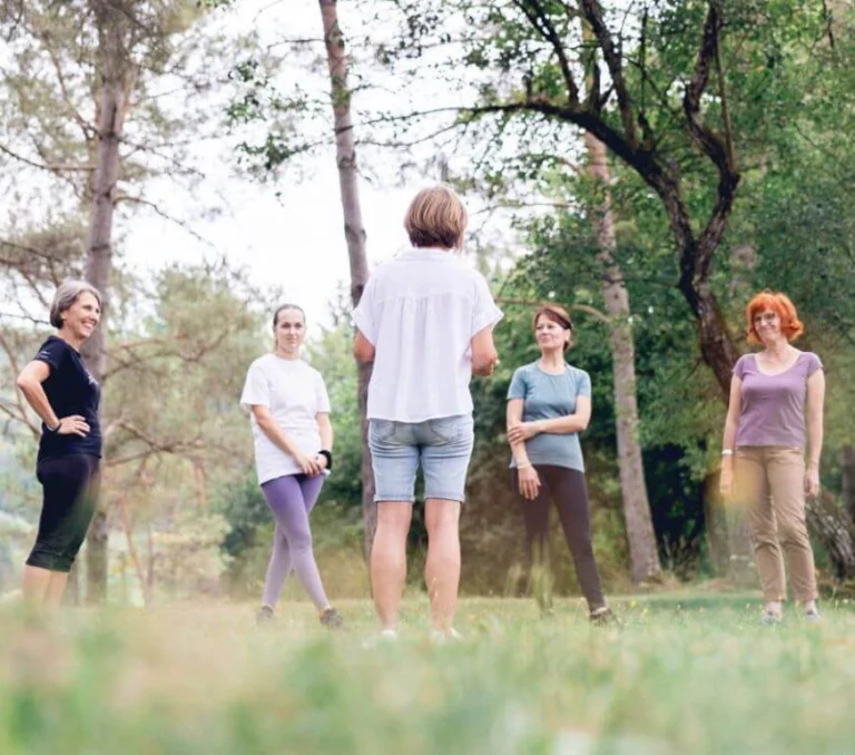 Au milieu des bois et avec une prise de vue au sol, Ilse Geusens est en train d’enseigner des techniques à un groupe de professionnels du bien-être qui se tient debout et en cercle.