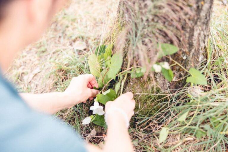 Avec ses deux mains, une participante est en train de créer un écorituel au pied d’arbre avec des éléments récupérés dans la nature : feuilles, fleurs et branches.