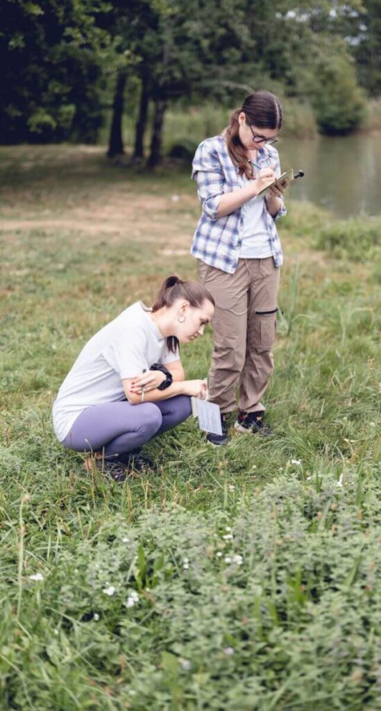 Dehors, tout près d’un lac, une naturopathe accroupie et une sophrologue debout sont en train de pratiquer un exercice enseigné lors de la formation d’écothérapie.
