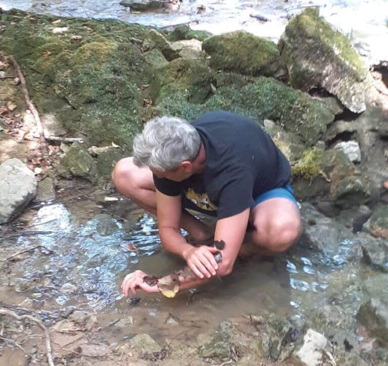 Un patient est accroupi dans un ruisseau et est en train de se libérer d’une émotion lourde en mettant des feuilles mouillées sur son bras gauche.