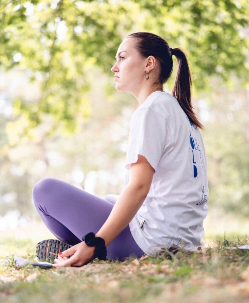 Sous un magnifique arbre, une prof de Yoga est assise en tailleur et observe la nature au loin avec les mains relâchées sur le sol.