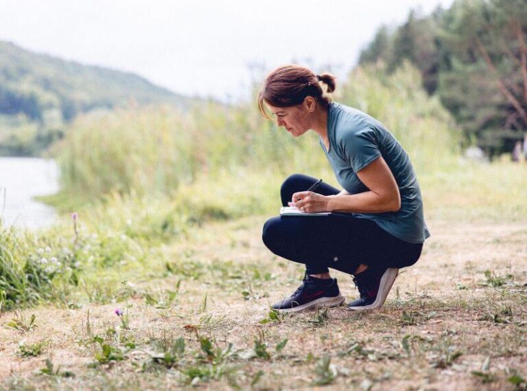 Accroupie et avec un cahier sur sa cuisse gauche, une sexologue est en train d’observer une fleur devant elle tout en mettant par écrit ses observations.