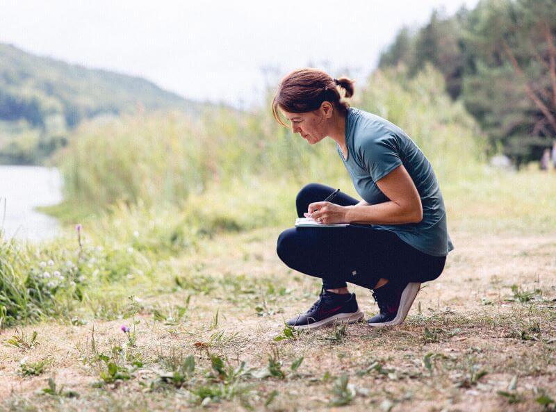Accroupie et avec un cahier sur sa cuisse gauche, une sexologue est en train d’observer une fleur devant elle tout en mettant par écrit ses observations.