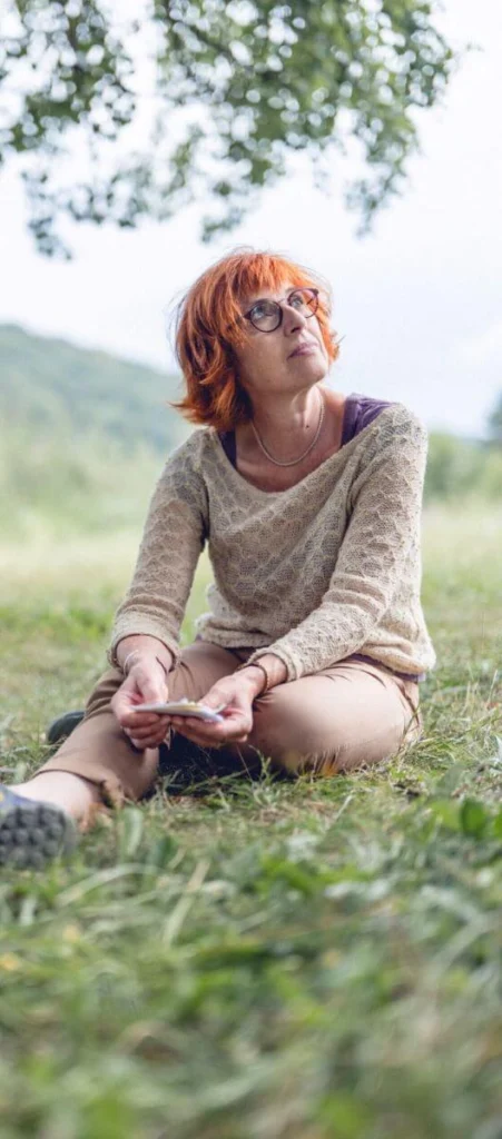 En pleine nature, une sophrologue est assise dans l’herbe et regarde en direction du ciel.