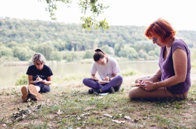 Tout près d’un lac, une accompagnatrice d’un collectif, une prof de yoga et une animatrice sont en train d’écrire sur leur carnet de notes.